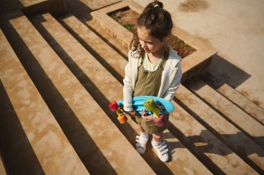 Kafkas görünümlü, günlük giysiler içinde küçük şirin bir kız çocuğu, yan bakıyor, elinde bir kaykayla merdivenlerde dikiliyor. Çocukluk, eğlence aktiviteleri. Aktif yaşam tarzı. Spor.