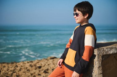 Yakışıklı, koyu renk saçlı, güneş gözlüğü ve spor giysisi takan, deniz manzarası karşısında güneşli bir günün tadını çıkaran genç bir çocuk. - İnsanlar. Yaşam tarzı. Boş zaman aktivitesi