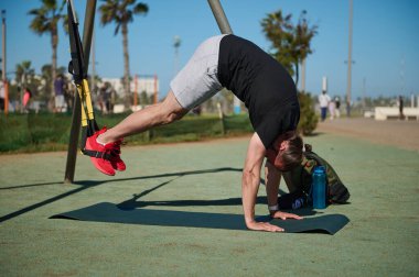 Direnç ve vücut ağırlığı egzersizleri yapıyor. Spor elbiseli genç bir atletik, açık hava spor sahasında, güneşli bir yaz gününde süspansiyon spor kemerleriyle bacaklarını çalıştırıyor. İnsanlar ve spor konsepti