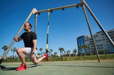 - İnsanlar. Spor. Aktif sağlıklı yaşam tarzı konsepti. Orta yaşlı erkek sporcu vücut ağırlığı egzersizleri yapıyor. Süspansiyon kayışlarını kullanıyor. Spor sahasında spor yaparken hamleler yapıyor.