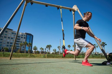 Egzersiz yaparken kaslı bir adam. Vücut ağırlığı ve süspansiyon kayışları. Atletik adam hamleler yapıyor, bacak kaslarını çalıştırıyor. Millet, aktif ve sağlıklı bir yaşam tarzı. Spor. Fitness