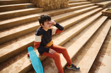 Genç bir öğrencinin portresi, spor giyimli okul çocuğu şehir parkındaki merdivenlerde oturuyor, düşünceli bir şekilde uzaklığa bakıyor. Yaz kaykayı, aktif yaşam tarzı. Spor eğlencesi.