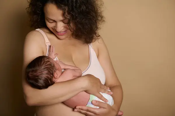 Mutlu genç anne, yeni doğmuş bebeğini kucağında tutarak olumlu duygular ifade ediyor, bej rengi stüdyo altyapısı üzerinde çıplak duruyor, doğumdan birkaç gün sonra doğum sonrası vücudunu kusurlu gösteriyor. Doğum Sonrası