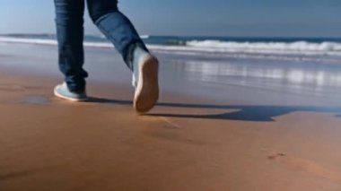 Islak sahil kumları ve ayak izleri. Güzel bir sahil boyunca yürürken mavi kot ayakkabılı kadın bacaklarının dikiz görüntüsü. Deniz suyu dalgaları kıyıdaki kumu dövüyor. Atlantik Okyanusu Fas