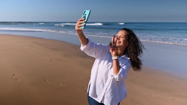 笑顔の幸せなカーリーハイレッド女性ブロガー笑顔 スマートフォンでビデオを録音し エアキスを送信し ビデオリンクで話し ソーシャルメディアで彼女のライフスタイルを共有し 海の砂浜に立つ — ストック動画