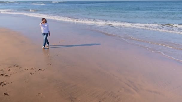身穿白衬衫和蓝色牛仔裤的多种族美女微笑着沿着海滨走着 在被海浪冲走的湿沙滩上留下了脚印 大西洋 大家好自然 — 图库视频影像