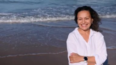 Beyaz gömlekli orta yaşlı güzel bir kadının yakın plan portresi, kolları deniz kıyısında katlanmış, sıcak bir sonbahar gününde güneşin tadını çıkarırken gülümsüyor. Dalgalar kıyıya vuruyor. İnsanlar ve doğa