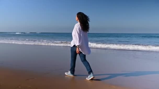 一个卷曲头发的女人穿着白衬衫和蓝色牛仔裤 享受她的户外假期 在温暖的阳光明媚的秋日沿着大西洋海岸跑步和散步的后视镜 人与美丽的自然 — 图库视频影像
