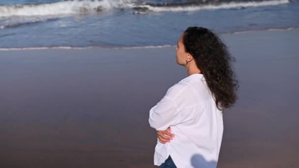 美丽的卷发拉丁黑发女人 穿着白衬衫 自信镇定 在阳光明媚的温暖天气里走在大西洋海滩上 享受她的户外周末 欣赏海浪拍岸 — 图库视频影像