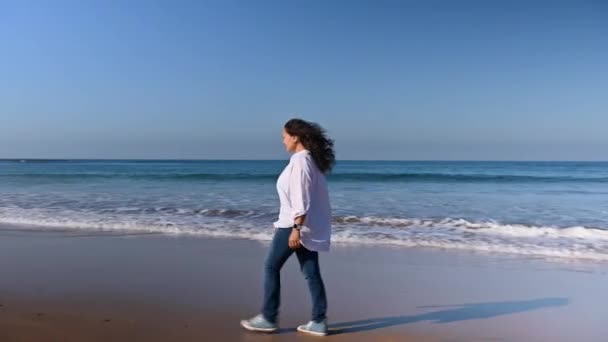 穿着蓝色牛仔裤 运动鞋和白衬衫的年轻快乐女人在大西洋沙滩上散步 在湿沙滩上留下脚印 浪涌上她的脚 — 图库视频影像