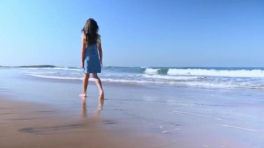Uzun saçlı, kot mayo giymiş, haftasonunun tadını çıkaran, kumsalda yalın ayak yürüyen, ıslak kumda dalgalarla yıkanan beyaz bir kızın arka görüntüsü..