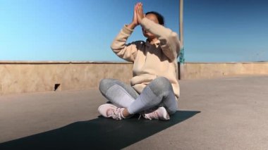Güzel Latin Amerikalı kadın nilüfer çiçeğiyle yoga yapıyor. Atlantik kıyısında poz veriyor, temiz hava soluyor, avuç içlerini bir arada tutuyor. Aktif, sağlıklı yaşam tarzı, boş zaman aktiviteleri, insanların hobisi