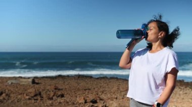 Şişeden su içen orta yaşlı, mutlu bir kadın. Yoğun bir idmandan sonra dinleniyor. Kameraya bakıp gülümsüyor.