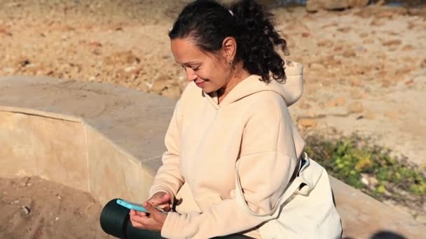 Χαμογελώντας Νεαρή Γυναίκα Πληκτρολογώντας Έξυπνο Τηλέφωνο Στην Παραλία Ελέγχοντας Περιεχόμενο — Αρχείο Βίντεο