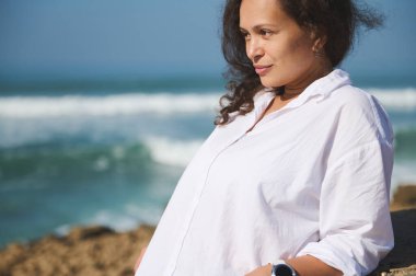 Kıvırcık saçlı, etnik kökenli genç bir esmerin yakın plan portresi, 35-40 yaşlarında güzel bir kadın, beyaz bir gömlek içinde, okyanus arka planına yaslanmış, rüya gibi uzaklığa bakıyor.