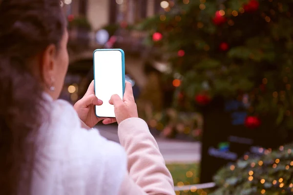 Close-up hands of a woman in winter clothes outdoors next to the Christmas tree, holding smart mobile phone with white blank mockup digital screen, copy ad space. Merry Christmas and New Year concept.