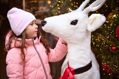 Sıcak kış kıyafetleri içinde güzel bir kız Noel ağacının yanında duran beyaz bir geyik oyuncağına hayran, çelenkler ve Noel ışıklarıyla aydınlatılmış, yaklaşan Noel tatilinin tadını çıkarıyor. Mutlu yıllar.