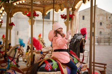 Mutlu kız gülümser ve atlıkarıncada eğlenirken el sallar Noel lunaparkında siyah madalyalı ata biner. İtalyan şehrinde Noel atmosferi Lombardiya 'da Como