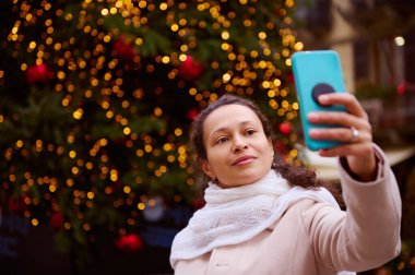 Çok ırklı güzel bir kadın akıllı telefonuyla selfie çekiyor, açık hava panayırında aydınlanmış Noel ağacına karşı duruyor, gülümsüyor ve olumlu duygularını ifade ediyor. İnsanlar ve iletişim