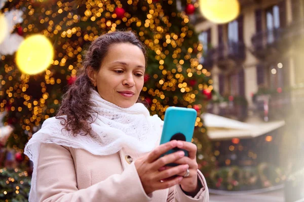 Close-up portrait of a multi ethnic beautiful young adult woman holding mobile phone, online shopping for presents, checking social media standing against illuminated Christmas tree with bokeh