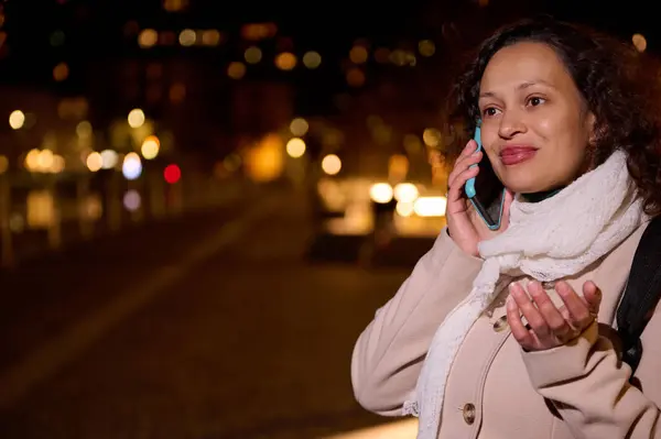Charming young adult multi ethnic woman smiling talking on mobile phone, standing on the street at night, with beautiful Christmas lights decorating the city at funfair. Copy advertising space