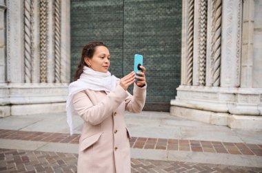 Orta yaşlı, şık bej ceketli muhteşem bir kadın, İtalya 'nın orta çağ şehri Como' yu ziyaret ediyor, elinde akıllı cep telefonuyla Santa Maria Maggiore katedralinin dış yüzünü fotoğraflıyor.