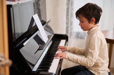 Yakışıklı bir çoklu etnik çocuğun otantik hayat tarzı portresi, piyano tuşlarına dokunmak, müzikal kompozisyon yaratmak ve piyanoda şarkı söylemek, kapalı alanda müzik dersi sırasında tek başına oturmak.