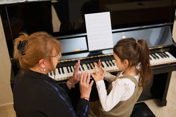 View from above of a female pianist teacher explaining piano lesson to a little child girl, showing the position of fingers on piano keys, enjoying together individual music lesson indoors