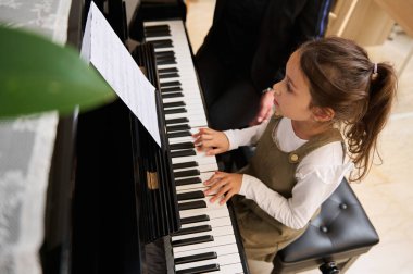 Piyano çalan küçük bir kızın en iyi görüntüsü, evde bireysel müzik dersi sırasında öğretmeninin rehberliğinde bir melodi bestelemesi. Çocuklar işte. Eğitim. Yaşam tarzları. Hobiler ve eğlence
