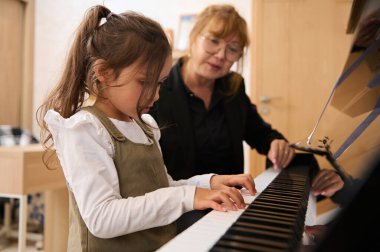 Olgun bir bayan müzisyen, müzik öğretmeni, piyanist, piyano öğretmeni, küçük bir kız öğrenciye piyano tuşlarındaki parmaklarının doğru pozisyonunu anlatıyor. - İnsanlar. Sanat, kültür ve eğlence.