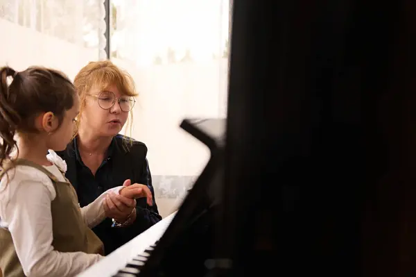 Caucasian middle aged female pianist musician teacher holding the hands of a cute student, showing her the the truth position of her finger on piano keys while performing classical musical composition