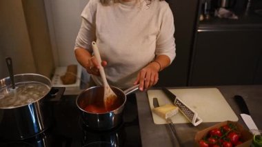 Ağır çekim. Genel olarak güzel bayan ev hanımı akşam yemeği için İtalyan makarnası pişiriyor, tencerede domates sosunu karıştırıyor akşamları sıcak ev mutfağında elektrikli sobanın yanında duruyor. Romantik ışıklandırma