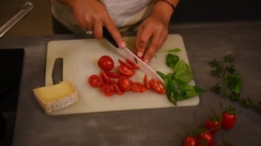 Üst Manzara. Ağır çekim. Kadın aşçı evde taze salata pişiriyor, beyaz kesim tahtasında organik sulu domatesleri dilimliyor. Mutfak masasında taze fesleğen yaprakları ve peynir. - İnsanlar. Sağlıklı gıda konsepti