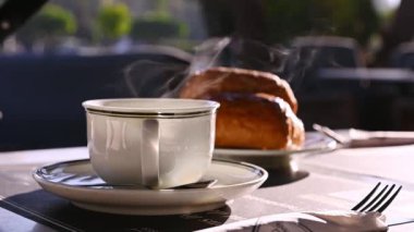 Durgun hayat. Ağır çekim. Bulanık Fransız kruvasanlarının arka planında buharlı beyaz bir fincan sıcak içecek, çay veya kahve amerikan kahvesi, açık bir kafede kahvaltıda servis edilir. Yiyecek ve içecek konsepti