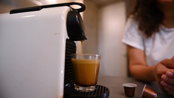 家庭用コーヒーマシンから注がれた新鮮なエスプレッソをクローズアップしたガラスカップ キッチンテーブルに立ってコーヒーを準備する輝かしい女性 新鮮なエスプレッソのカップで一日を始める — ストック動画
