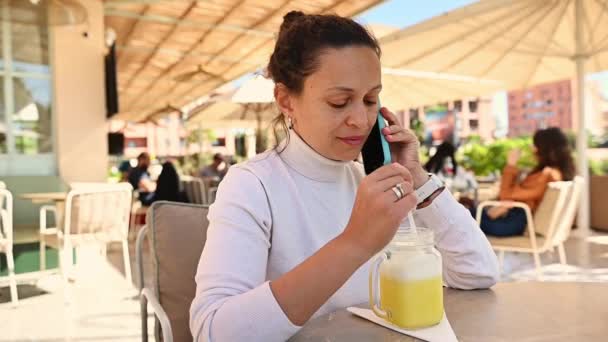 一个中年妇女在手机上说着话 微笑着 在户外咖啡馆里喝着新鲜的果汁 慢动作 稳稳地拍了下来 — 图库视频影像