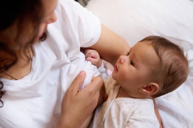Beyaz elbiseli, beyaz elbiseli, annesinin onu emzirirken ve yatakta yan yana otururken ona gülümseyen beyaz erkek bebeğin yakın plan portresi. Bebek ve annelik izni kavramı