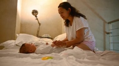 Genç bir anne, 4-6 aylık bebeğiyle hijyenik bezini değiştirip rahat yatak odasında yatakta oturuyor. Bebek bakımı, bebeklik ve bebeklik konsepti. Annelik ve annelik. - İnsanlar. Yaşam biçimi