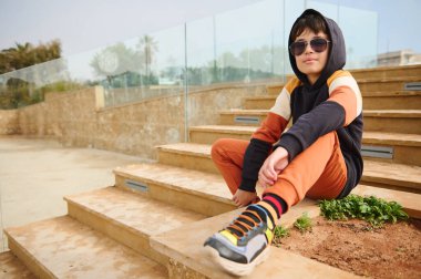Yakışıklı, şık spor kıyafetleri ve güneş gözlüklü genç bir çocuk, kameraya özgüvenle bakıyor, merdivenlerde oturuyor, dışarıda mutlu bir hafta sonunun tadını çıkarıyor. İnsanlar ve yaşam tarzları. Reklam alanını kopyala