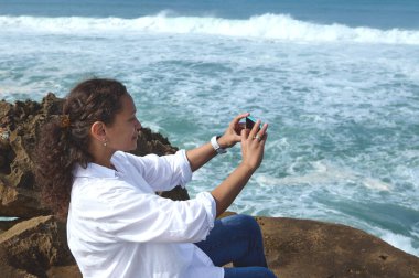 Kıvırcık saçlı, çok ırklı güzel bir kadın akıllı cep telefonuyla fotoğraf çekiyor Atlantik Okyanusu sahilindeki kayanın üzerinde oturuyor. İnsanlar ve doğa. Doğayla bağlantı. Eğlence için. Boş zaman aktivitesi