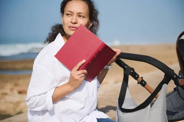 Beautiful woman holding a hard covered book near her face, dreamily looking away, sitting on the parapet on the beach and pushing baby pram, against Atlantic ocean background. People. lifestyles