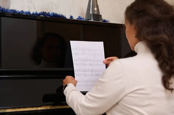 Rear view of female pianist holding notes of classic musical composition, sitting at black old piano, ready to play a melody. Art, culture and entertainment.