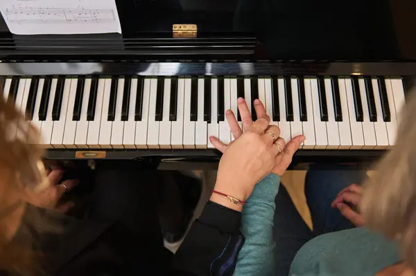 View from above women holding hands on piano ebony and white keys, performing classical musical composition, feeling rhythm while play grand piano at home. Music lessons for adults. People. Education