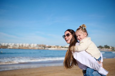 Genç beyaz bir kadının yan portresi, sevgi dolu bir anne güzel kızına kucak açıyor, birlikte kumsalda mutlu anlar geçiriyor, geniş bir gülümsemeyle ve hayallerle uzaklığa bakıyor.