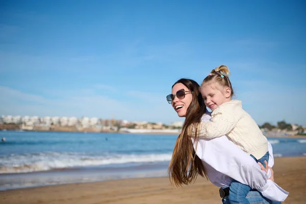 Side portrait of a young Caucasian woman, loving mother giving piggyback to her lovely child girl, spending happy moments together on the beach, smiling broadly and dreamily looking into the distance