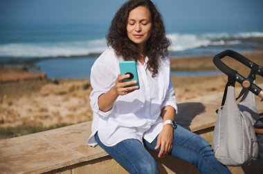 Mutlu bir anne cep telefonu kullanıyor, kumsalda tatlı çocuğuyla oturuyor, bebek arabasında uyuyor.