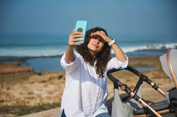 Happy young mom smiles broadly while takes selfie on her modern smart phone, walking with her baby sleeping in baby carriage, sitting on parapet against Atlantic ocean shore. People. Lifestyle. Nature