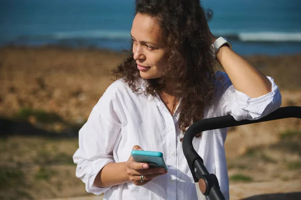 Portrait of a pretty woman of Latin American ethnicity holding smart mobile phone, dreamily looking into the distance, sitting against sea background. Young mom using mobile phone outdoor