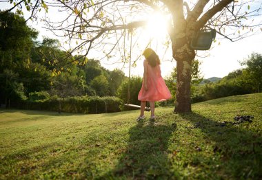 Gün batımında büyük yeşil bir bahçede salıncakta sallanan salıncağın yanında duran pembe elbiseli, güzel, beyaz bir kız çocuğu. Tam boy portre. - İnsanlar. Çocukluk. Boş zaman aktivitesi