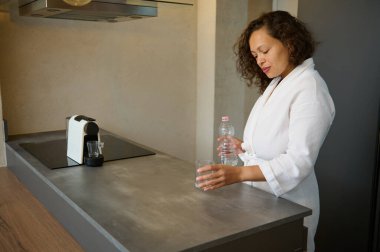 Beyaz waffle bornozlu otantik orta yaşlı kadın güne bir bardak sulandırıcı tatlı suyla başlıyor. Sağlık ve vücut bakımı konsepti. Sağlıklı bir yaşam tarzı. Sabah rutini ve alışkanlıkları